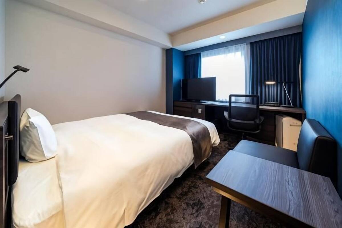 Una habitación de hotel con cama, escritorio, silla, televisión y lámpara.