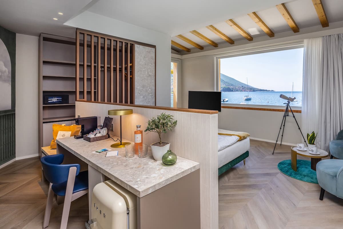 Een moderne hotelkamer met een houten plafond, een groot raam met uitzicht op zee, een bureau met lamp, een stoel, een bed en een telescoop op een statief.