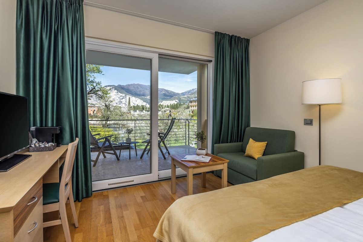 Hotelkamer met een bed, een bureau met een computer, een groene bank, een tafeltje en uitzicht op bergen door een glazen schuifdeur.