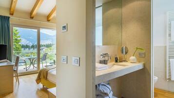 A modern hotel room with a large window, a balcony, a wooden ceiling, a bathroom with a large mirror, and a bed.