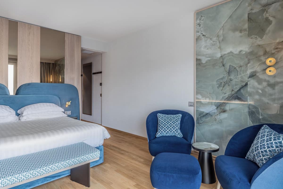 Een moderne hotelkamer met een groot bed, twee blauwe fauteuils, een klein bijzettafeltje en een marmeren wand.