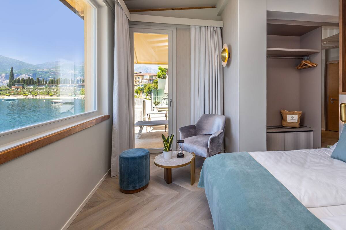 Hotelkamer met een bed, een stoel, een klein tafeltje met een plant, en uitzicht op water en gebouwen.