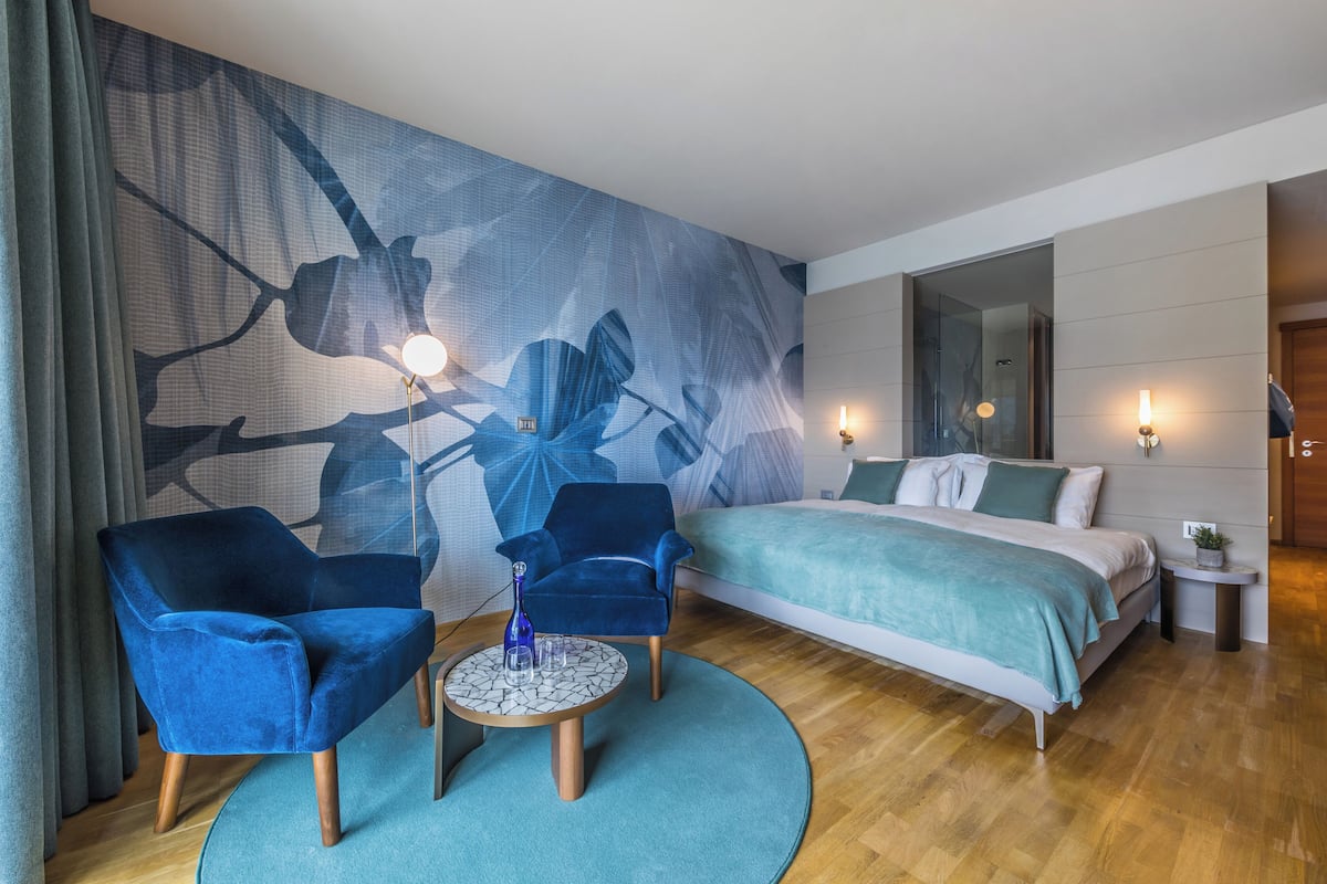 Een moderne hotelkamer met een groot bed, twee blauwe fauteuils, een klein rond tafeltje en een groot abstract schilderij aan de muur.
