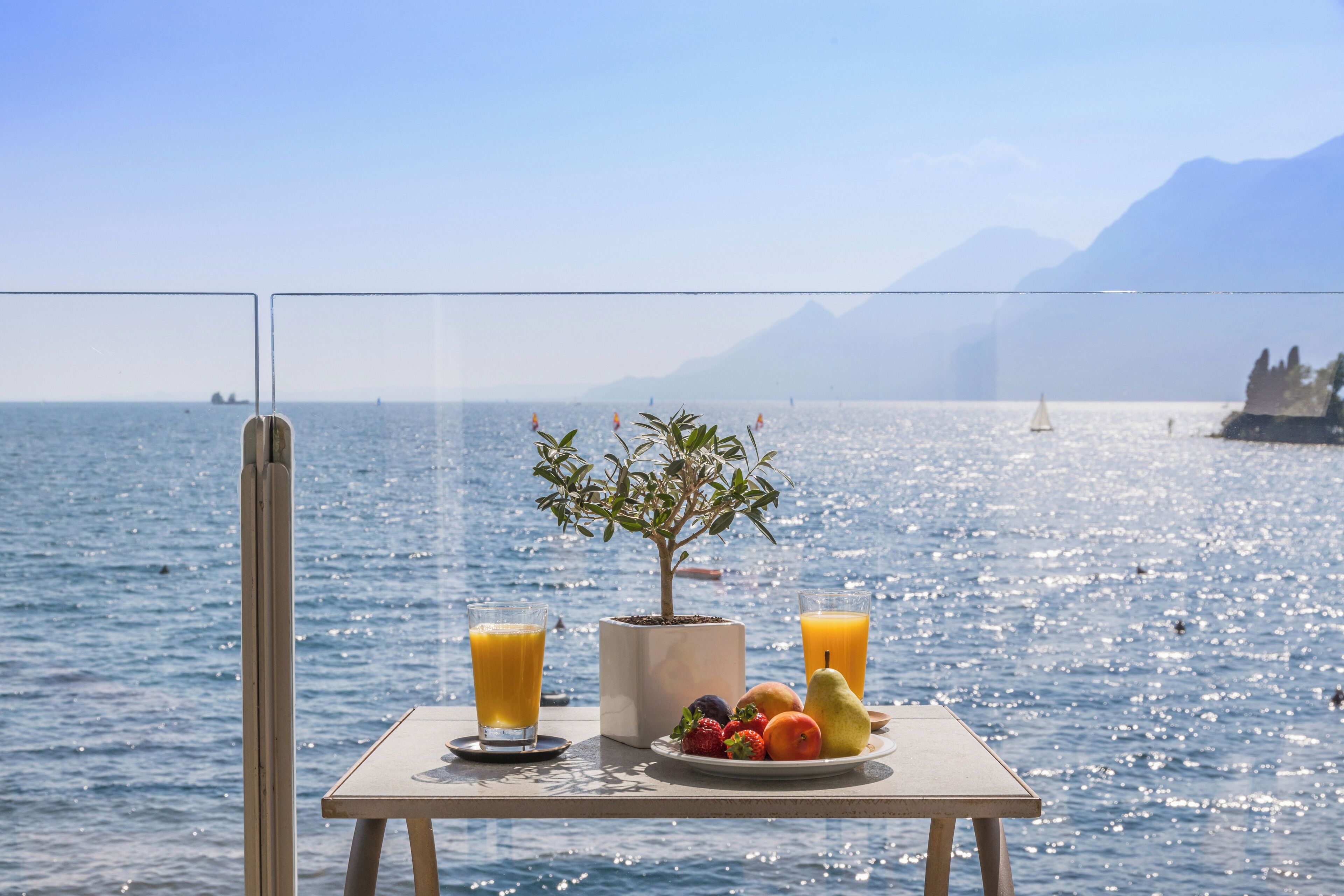 Une table avec des fruits et des verres de jus, placée au bord d’un lac pittoresque, avec des montagnes en arrière-plan.