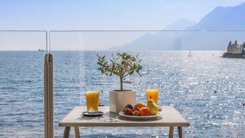 Une table avec des fruits et des verres de jus, placée au bord d’un lac pittoresque, avec des montagnes en arrière-plan.