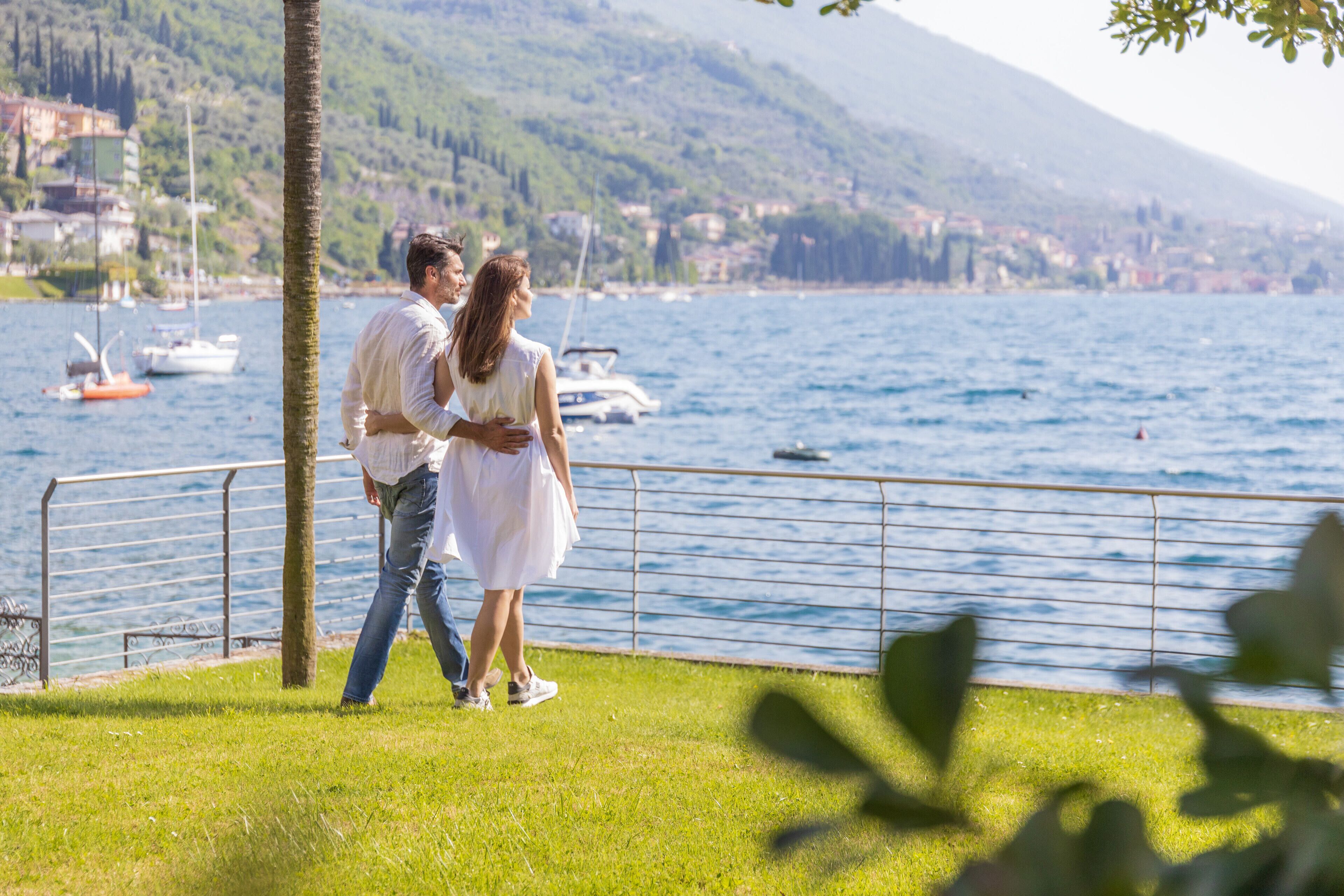 Una pareja pasea por un paseo marítimo junto al lago, con vistas a barcos y montañas al fondo.