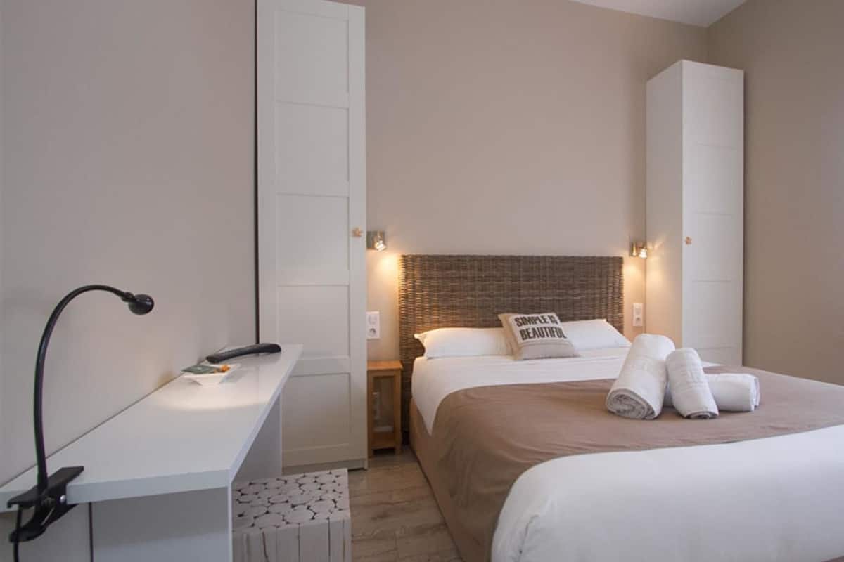 Quarto de hotel com cama, escrivaninha, mesa de cabeceira e janela.
