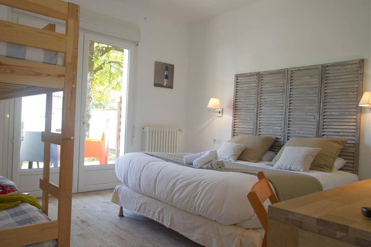 Quarto com beliche, uma cama de solteiro, escrivaninha de madeira e vista para o exterior.