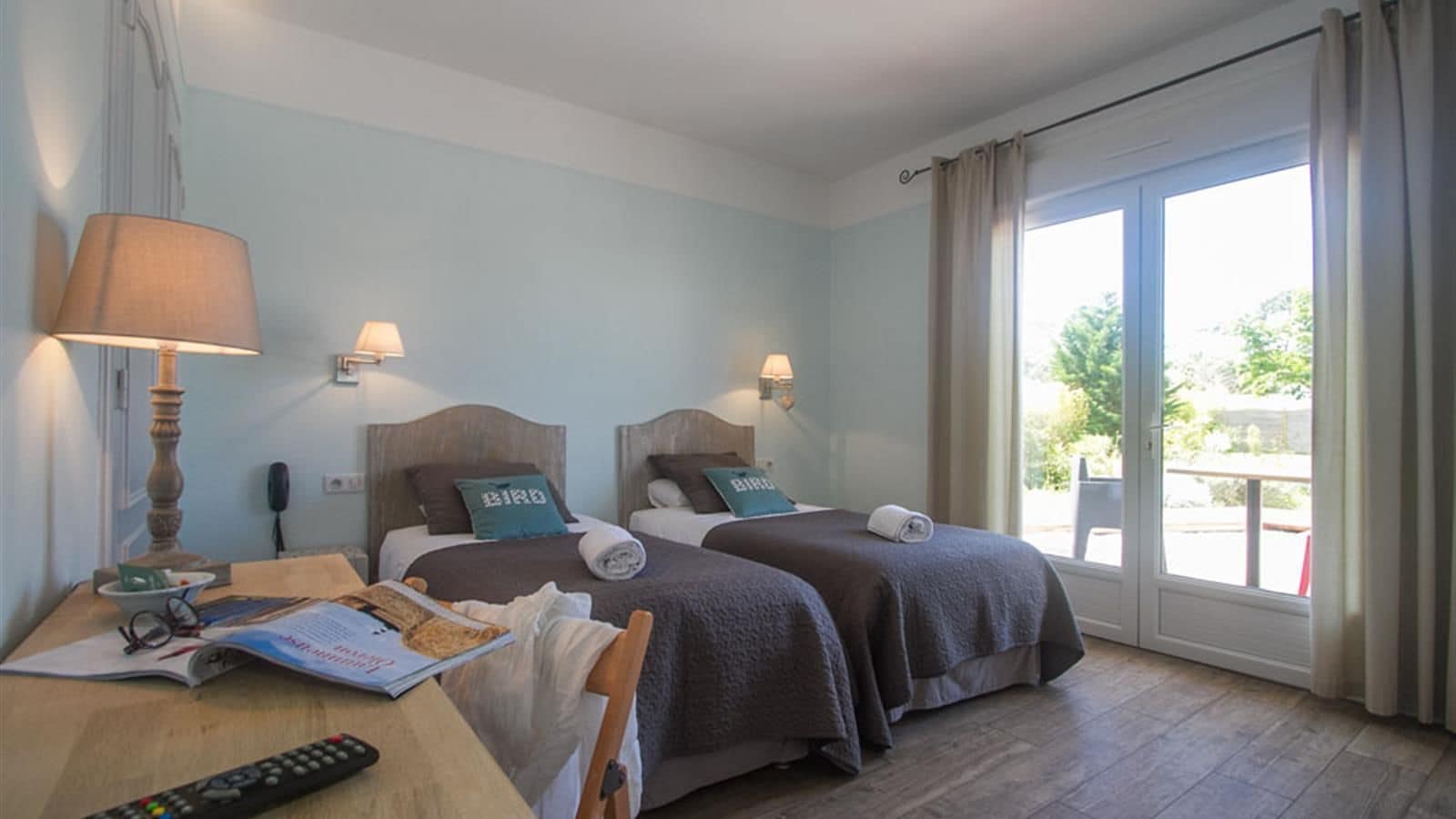 A hotel room with two beds, a desk with a lamp, a remote control, and a window with curtains.