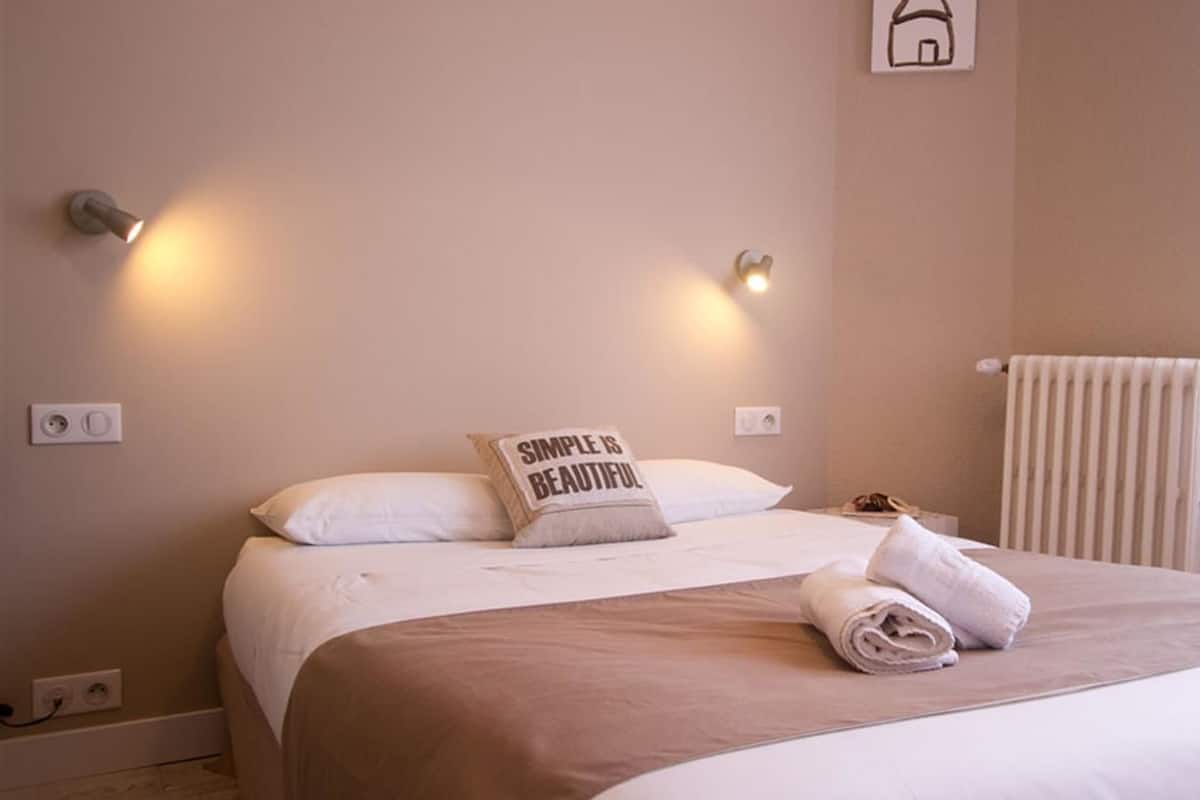 Uma cama bem arrumada com um travesseiro que diz "Simple is Beautiful", um candeeiro fixado na parede e um radiador.