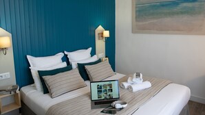 Een net opgemaakt bed met een laptop, een koptelefoon en een opgerolde handdoek in een hotelkamer met een blauwgroen hoofdeinde en een schilderij van een strand aan de muur.