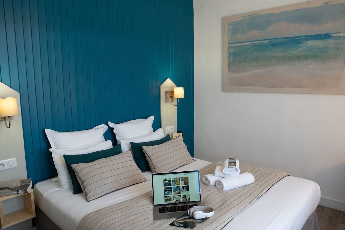 Cama bem arrumada com um laptop, um par de fones e uma toalha enrolada em quarto de hotel com cabeceira azul-turquesa e um quadro de praia na parede.