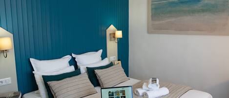 A neatly made bed with a laptop, a pair of headphones, and a rolled towel on a hotel room with a teal headboard and a painting of a beach on the wall.