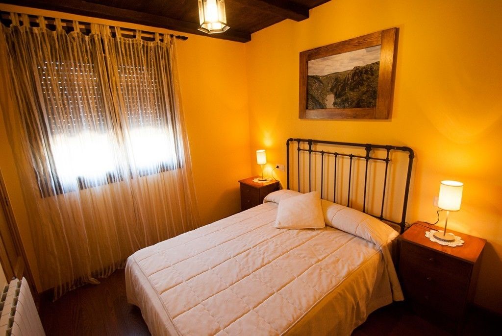 Un dormitorio con cama, dos mesitas de noche, una lámpara, una ventana con cortinas y un cuadro en la pared.
