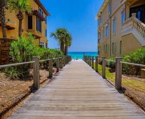 A wooden boardwalk leading to a beach with clear blue water, flanked by buildings with balconies and palm trees. - Casa de Fiesta 5 Bedroom Holiday Home by Five Star Properties (Destin)