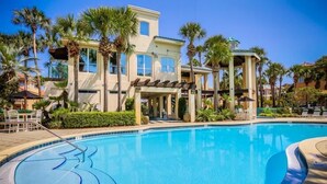 A swimming pool with a curved edge, surrounded by palm trees and a building with a light-colored facade and multiple windows. - Casa de Fiesta 5 Bedroom Holiday Home by Five Star Properties (Destin)