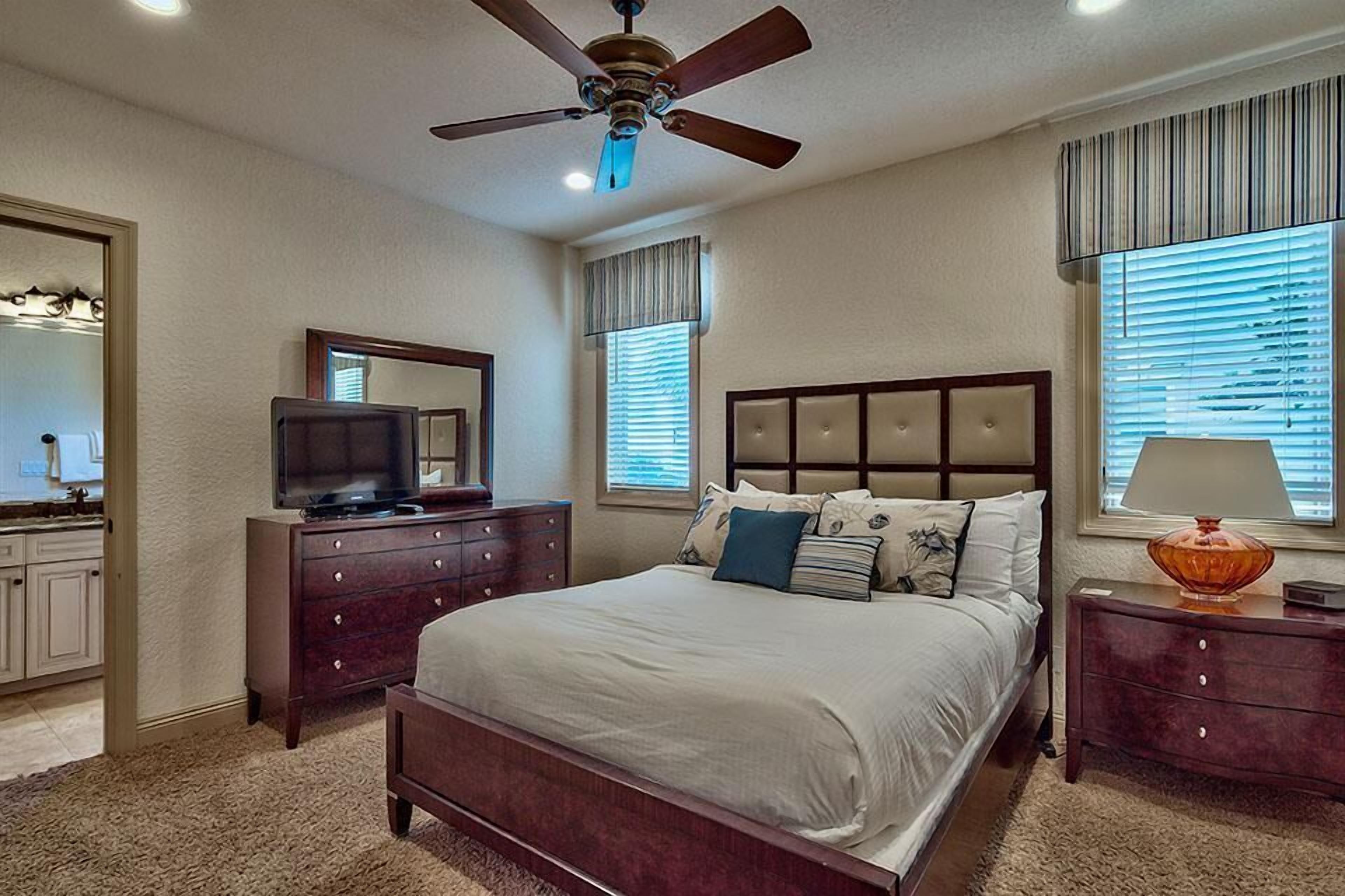 A bedroom with a bed, a dresser, a television, a ceiling fan, and windows with blinds.