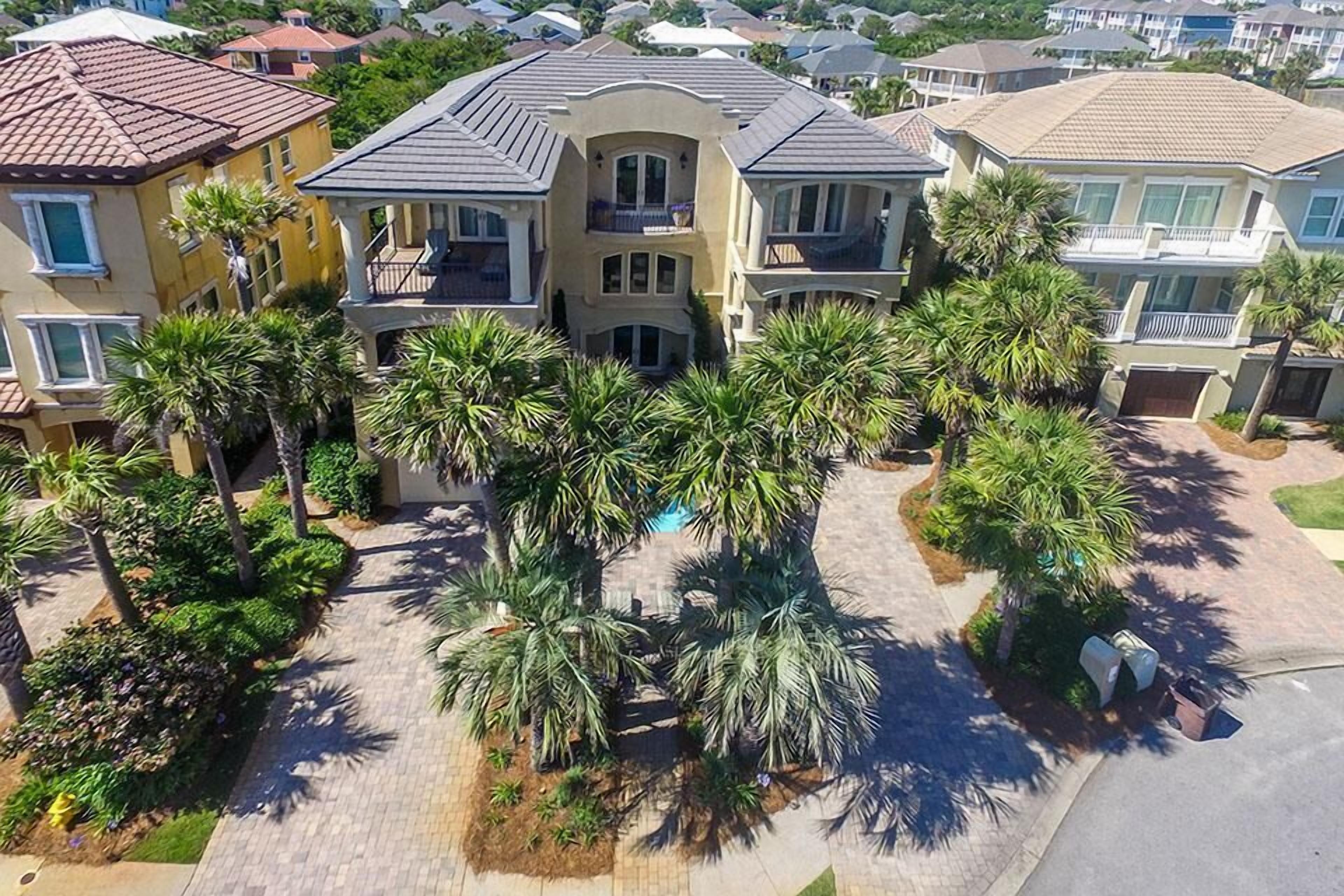 A multi-story house with a balcony, surrounded by palm trees and a paved driveway.