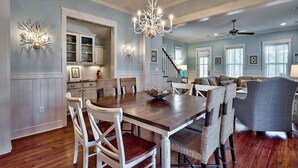 Un vaste espace dédié à la salle à manger, avec une table en bois et des chaises blanches, un lustre et un escalier menant à un autre niveau.