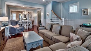 Un salon avec un canapé, une table basse et un fauteuil. On y trouve un escalier avec une rampe et une fenêtre avec des stores.
