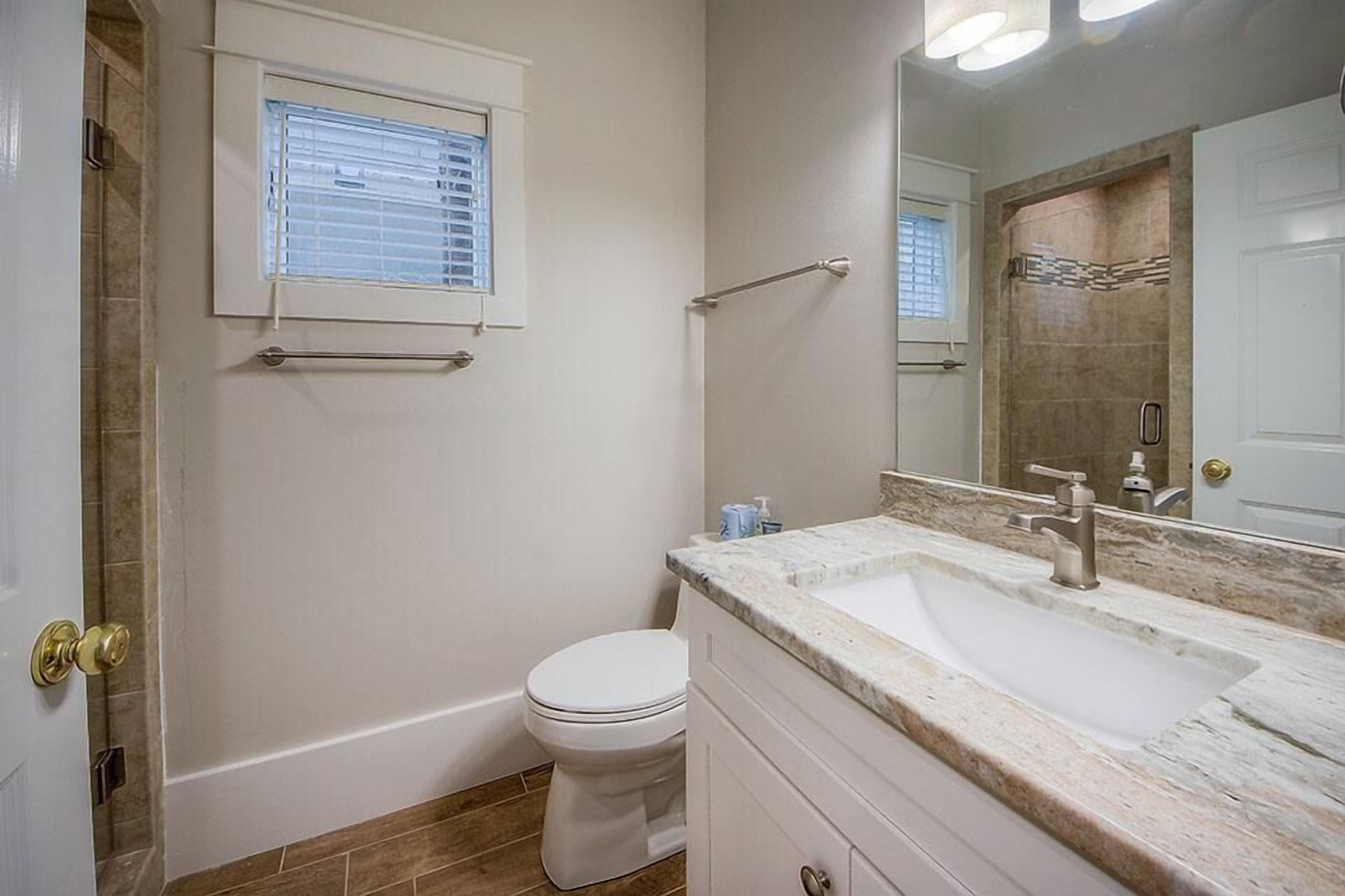 Une salle de bain avec des toilettes blanches, un lavabo doté d’un plan de travail en marbre, un porte-serviettes et une fenêtre avec des stores.