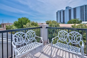 Ein Balkon mit zwei weißen Metallbänken und Geländer, mit Blick auf eine Stadtlandschaft mit hohen Gebäuden und Grünflächen.