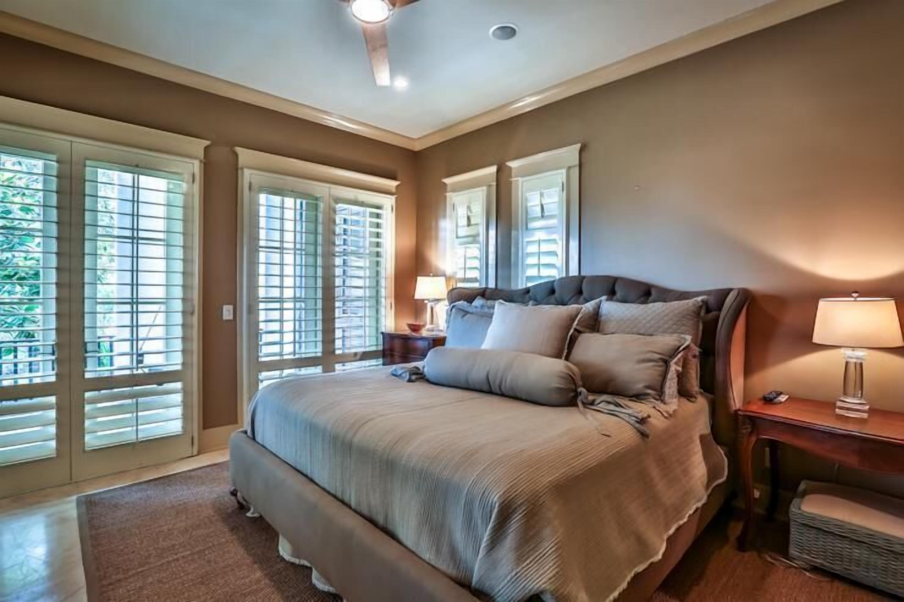 A bedroom with a large bed, a nightstand, a lamp, and a view through the window blinds.