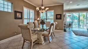 A dining area with a glass-top table, four upholstered chairs, a chandelier, and large windows with blinds. - Emerald Enchantment 4 Bedroom Holiday Home by Five Star Properties (Destin)