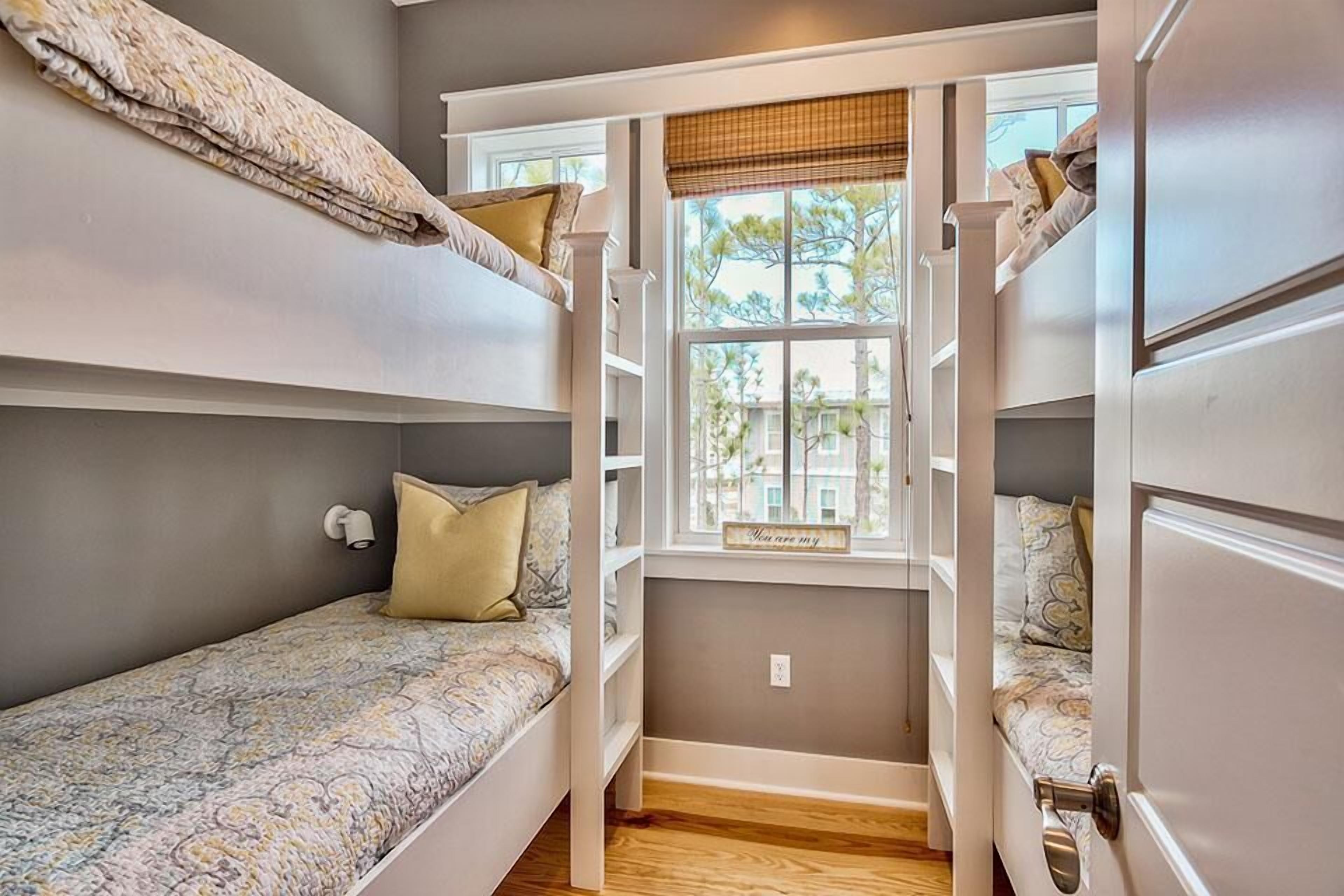 A dorm-style room with bunk beds, a window with a view of trees, and a small shelf with a sign.