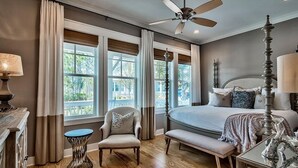 Quarto com cama, cadeira, mesa, ventoinha no teto e grandes janelas.