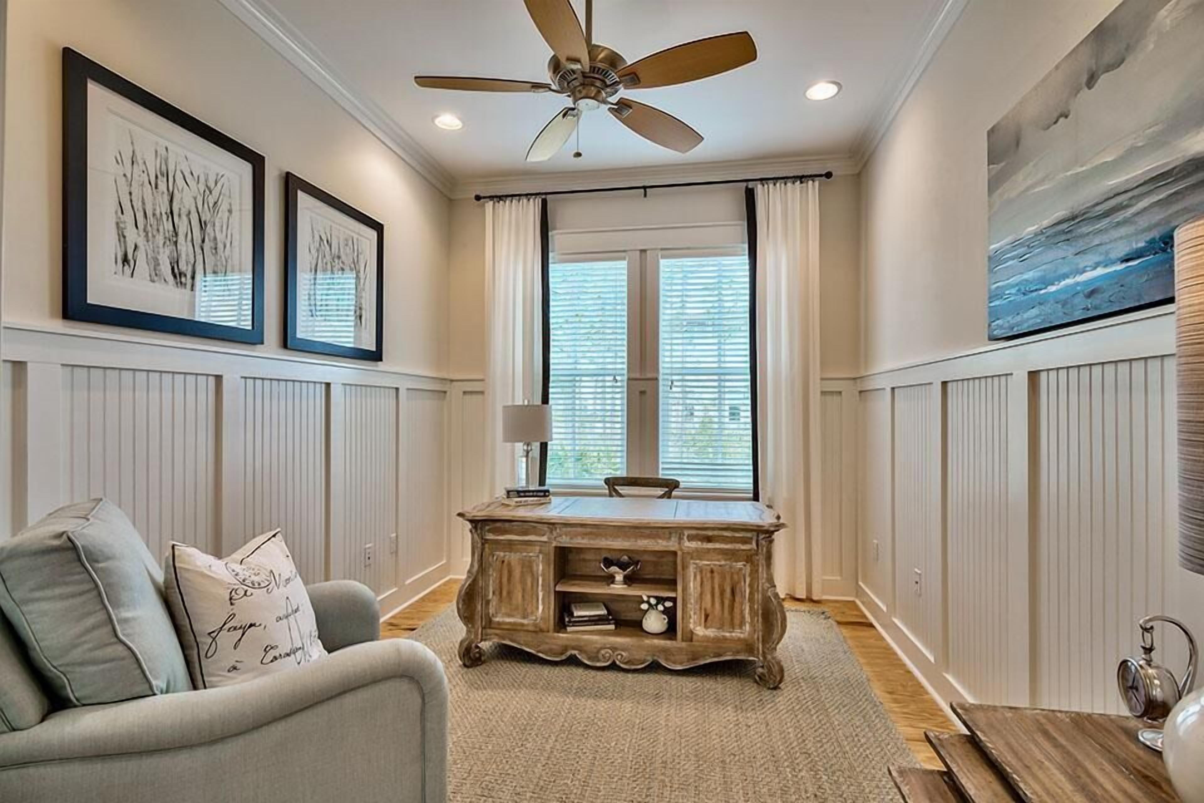 A room with a ceiling fan, a desk with a lamp, a chair, and framed artwork on the walls.