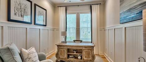 A room with a ceiling fan, a desk with a lamp, a chair, and framed artwork on the walls.