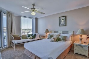 Une chambre d’hôtel avec un grand lit, deux tables de chevet avec des lampes, un ventilateur de plafond, une fenêtre avec des stores et un balcon avec une chaise.