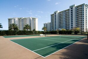 Ein Tennisplatz mit umgebender Grünfläche und Hochhäusern im Hintergrund.