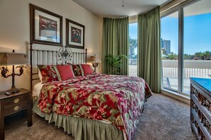 A hotel room with a large bed, two framed pictures on the wall, a nightstand with a lamp, a potted plant, and a view of the outside balcony.