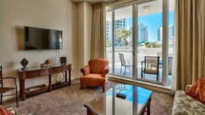 Ein Hotelzimmer mit Flachbildfernseher, einem Glastisch, einer Couch, einem Sessel und einem Balkon mit Sitzgelegenheit.