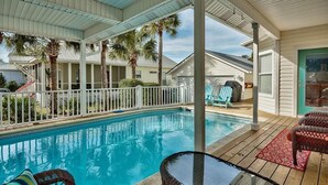 A pool area with a covered deck, lounge chairs, and a glass table.