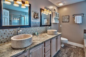 Un baño con dos lavamanos redondos, un espejo grande y un inodoro. Hay luces decorativas sobre los lavamanos y un perchero en la pared.