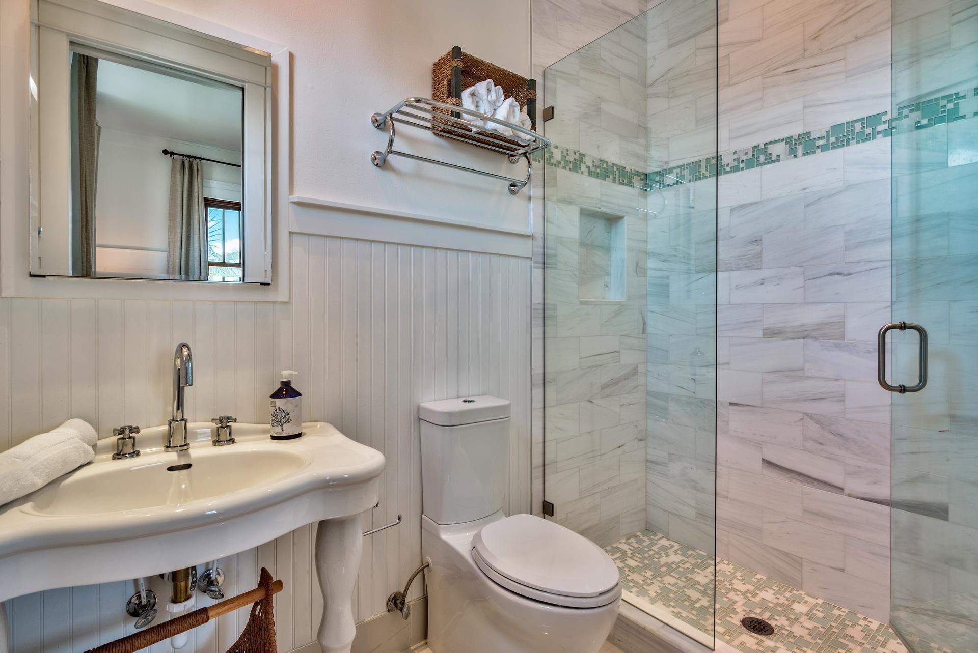 A bathroom with a white sink, mirror, and toilet. There is a glass shower enclosure with a tiled floor.