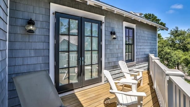 A wooden deck with two white Adirondack chairs and a small table, a sliding glass door, and a window with a view of the surrounding trees. - The Flagship of Watersound 5 Bedroom Holiday Home by Five Star Properties (Santa Rosa Beach)