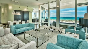 Un salon moderne doté d’une grande fenêtre donnant sur une plage, ainsi que d’un coin repas et d’une cuisine.