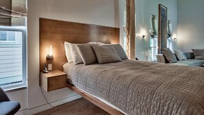 Un dormitorio moderno con cabecera de madera, mesita de noche con lámpara y vistas a la ciudad a través de grandes ventanales.