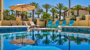 A pool area with lounge chairs and umbrellas, surrounded by palm trees and a building with a tiled roof. - Tuscan Villa 5 Bedroom Holiday Home by Five Star Properties (Destin)