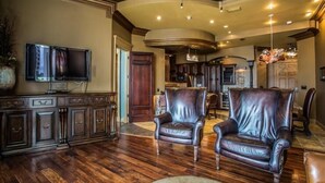 A spacious living room with dark wood flooring, leather armchairs, a flat-screen TV, and a kitchen in the background.
