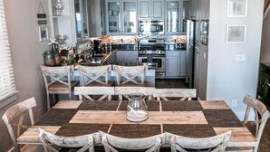 A dining area with a wooden table and chairs, a kitchen in the background, and a window with blinds. - Ventanas al Paraiso 5 Bedroom Holiday Home by Five Star Properties (Miramar Beach)