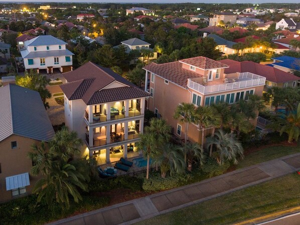 A multi-story residential building with a balcony, surrounded by palm trees and other houses.