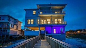 A multi-story beachfront house with a wooden walkway leading to the entrance.