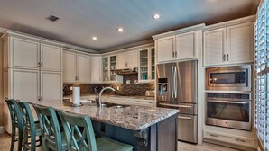 A modern kitchen with white cabinetry, a granite island, stainless steel appliances, and teal bar stools. - Honu Hale 5 Bedroom Holiday Home by Five Star Properties (Destin)