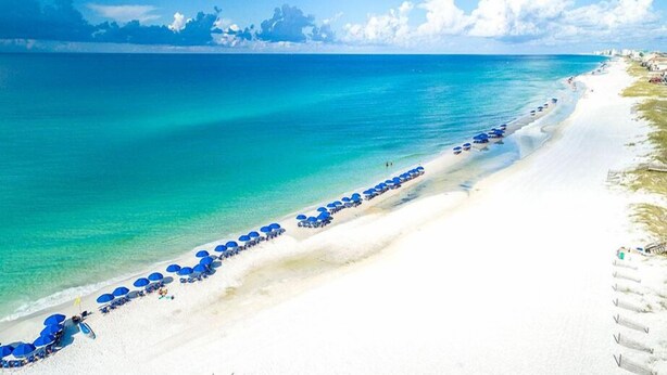 Une plage de sable blanc bordée d’une eau turquoise et cristalline. De nombreux parasols bleus et des chaises de plage sont alignés le long du rivage.