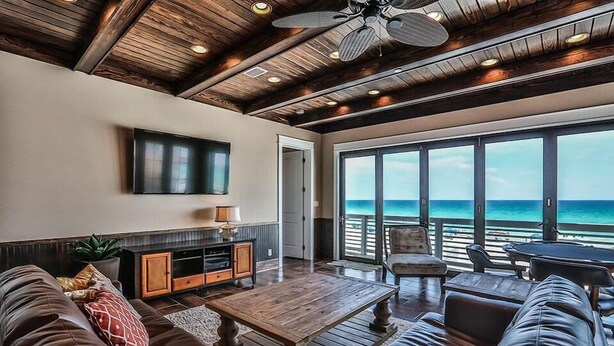 A spacious living area with a wooden ceiling, a large TV mounted on the wall, and a view of the ocean through large windows. - Kates Porch 7 Bedroom Holiday Home by Five Star Properties (Miramar Beach)